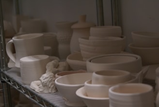 A collection of unfinished ceramic pieces, including mugs, bowls, and containers, is neatly arranged on metal shelving. The pieces are mostly unglazed and have a matte, neutral color, suggesting they are in the early stages of pottery making.