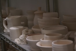 A collection of unfinished ceramic pieces, including mugs, bowls, and containers, is neatly arranged on metal shelving. The pieces are mostly unglazed and have a matte, neutral color, suggesting they are in the early stages of pottery making.