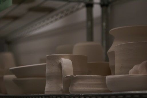 Several pieces of unglazed pottery are arranged on a metal shelf. The pottery includes bowls, cups, and vases, all with a smooth, light beige surface. The lighting is dim, creating a soft shadow across the objects, which appear to be works in progress or ready for further finishing.