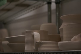 Several pieces of unglazed pottery are arranged on a metal shelf. The pottery includes bowls, cups, and vases, all with a smooth, light beige surface. The lighting is dim, creating a soft shadow across the objects, which appear to be works in progress or ready for further finishing.