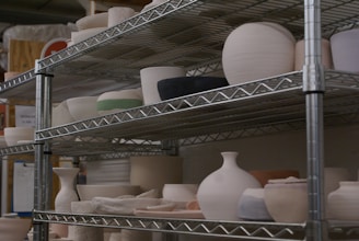 Metal shelves filled with various pottery items, including bowls, vases, and pots in different shapes and sizes. The pottery appears to be unfinished or in different stages of completion, with a predominantly matte finish.