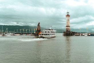 A large boat with a German flag sails past a lighthouse and a statue of a lion at the entrance of a harbor. The scene is set under an overcast sky with mountains visible in the background. Several smaller boats are docked nearby, and yellow flags adorn the jetty.