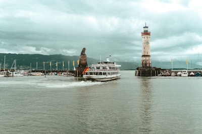 A large boat with a German flag sails past a lighthouse and a statue of a lion at the entrance of a harbor. The scene is set under an overcast sky with mountains visible in the background. Several smaller boats are docked nearby, and yellow flags adorn the jetty.