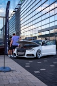 A white car is displayed with its hood open in an urban setting. Several people are gathered around, examining or discussing the vehicle. The backdrop features a modern glass building that reflects a blue sky, and a tall flag with the word 'Autocult' is visible.
