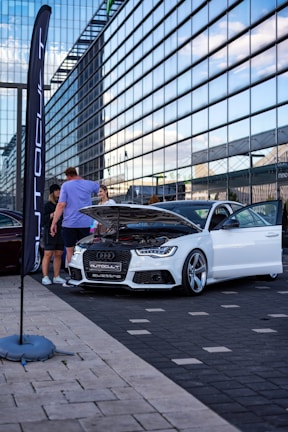 A white car is displayed with its hood open in an urban setting. Several people are gathered around, examining or discussing the vehicle. The backdrop features a modern glass building that reflects a blue sky, and a tall flag with the word 'Autocult' is visible.