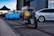A motorcycle with a yellow gas tank is parked on a paved parking area. Behind it, there are two cars, one blue and the other silver. A tent is set up nearby, under which a few people are standing. The background features a tall, glass-fronted building reflecting the sky.