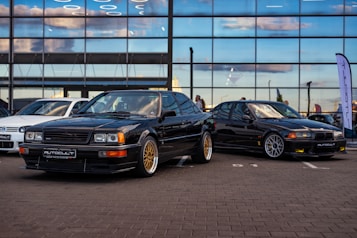 Two sleek, black vintage cars with sporty designs are prominently displayed in front of a large building with reflective glass windows. The cars are parked on a paved surface, with one having gold rims and the other featuring silver. A white car is visible in the background, and there is a tall flag or banner on the side. The reflections on the windows show a partly cloudy sky.