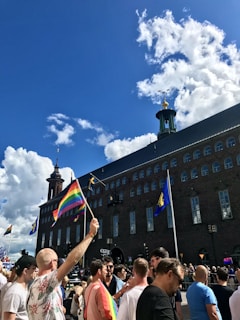 A crowd of people gathered in front of a large historic building, holding rainbow and Swedish flags. The sky is clear with a few clouds, and the mood appears festive and celebratory.