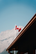 A Canadian flag waves prominently with a backdrop of majestic mountains. The sky is clear and blue, creating a serene and picturesque scene. The roof of a building can be seen in the bottom foreground, adding depth to the composition.