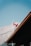 A Canadian flag waves prominently with a backdrop of majestic mountains. The sky is clear and blue, creating a serene and picturesque scene. The roof of a building can be seen in the bottom foreground, adding depth to the composition.