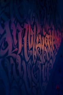 Intricate and stylized graffiti covers a wall, featuring a blend of swirling and interconnected shapes and characters. The art is accentuated by moody lighting that creates a gradient of dark blues transitioning into soft oranges, highlighting the texture and complexity of the design.