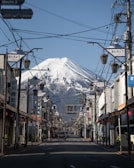 A scenic street with traditional Japanese architecture leads towards a spectacular view of a snow-capped mountain in the background. Street lamps and power lines frame the road, while several storefronts featuring Japanese signage line both sides. A car is visible on the road, and the sky is clear and blue.