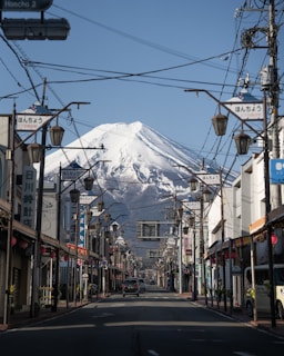 A scenic street with traditional Japanese architecture leads towards a spectacular view of a snow-capped mountain in the background. Street lamps and power lines frame the road, while several storefronts featuring Japanese signage line both sides. A car is visible on the road, and the sky is clear and blue.