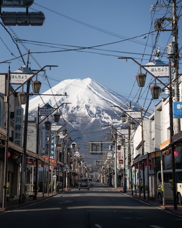 A scenic street with traditional Japanese architecture leads towards a spectacular view of a snow-capped mountain in the background. Street lamps and power lines frame the road, while several storefronts featuring Japanese signage line both sides. A car is visible on the road, and the sky is clear and blue.