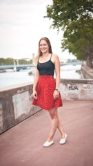 A young woman stands on a paved walkway next to a river. She wears a black top and a red skirt with patterns, along with white shoes. The background shows a tree-lined path and a body of water, possibly a river, with a bridge in the distance. The concrete wall beside her is covered with graffiti.
