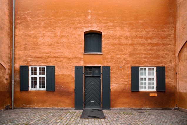 The image features an exterior view of a building with a textured orange wall. There is a green door centrally located, flanked by two windows with matching green shutters. The windows are white-framed with multiple panes. The ground is made of cobblestones, and there is a small rectangular plaque on the right side of the wall.