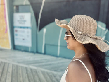Giovana walking along a sunlit Palm Beach boardwalk wearing a flowing white dress and oversized sunglasses.
