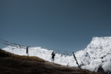 Snow-covered mountains rise majestically against a clear blue sky, with a person standing on a grassy hilltop. Colorful prayer flags are strung between poles, adding a spiritual or cultural element to the scene.