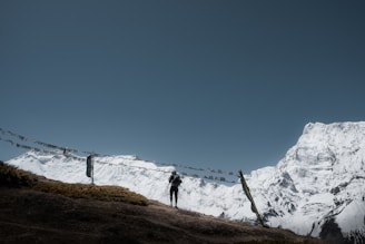 Snow-covered mountains rise majestically against a clear blue sky, with a person standing on a grassy hilltop. Colorful prayer flags are strung between poles, adding a spiritual or cultural element to the scene.
