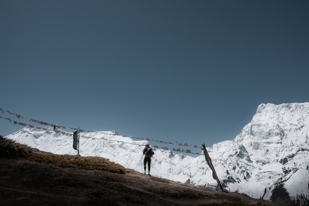 Snow-covered mountains rise majestically against a clear blue sky, with a person standing on a grassy hilltop. Colorful prayer flags are strung between poles, adding a spiritual or cultural element to the scene.