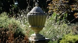 A large stone urn is set in a lush garden, surrounded by various shrubs and wildflowers. The urn has intricate detailing and a pointed lid. Sunlight casts shadows, highlighting the textures and colors of the foliage.
