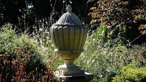A large stone urn is set in a lush garden, surrounded by various shrubs and wildflowers. The urn has intricate detailing and a pointed lid. Sunlight casts shadows, highlighting the textures and colors of the foliage.