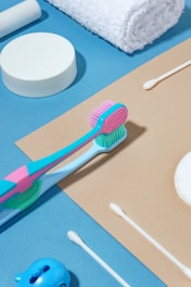 Various toiletries are arranged neatly on a blue and beige background, including a colorful toothbrush, cotton swabs, a white rolled towel, and a round container.