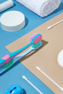 Various toiletries are arranged neatly on a blue and beige background, including a colorful toothbrush, cotton swabs, a white rolled towel, and a round container.
