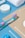 Various toiletries are arranged neatly on a blue and beige background, including a colorful toothbrush, cotton swabs, a white rolled towel, and a round container.