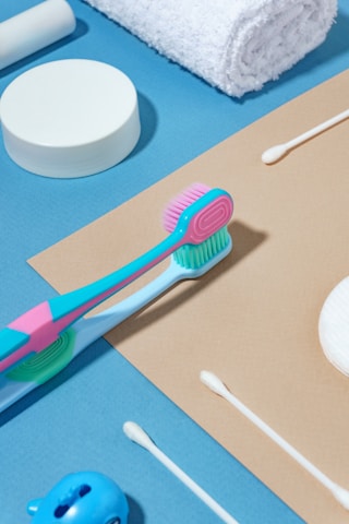 Various toiletries are arranged neatly on a blue and beige background, including a colorful toothbrush, cotton swabs, a white rolled towel, and a round container.