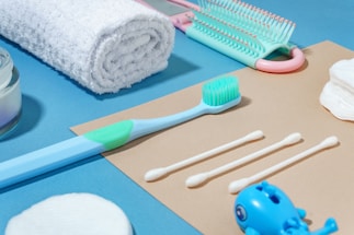 A neatly arranged collection of personal hygiene items on a two-toned background, including a rolled white towel, a toothbrush, a set of cotton swabs, a hairbrush, cotton rounds, and a small blue toy. The arrangement is aesthetically pleasing and conveys a sense of cleanliness and order.