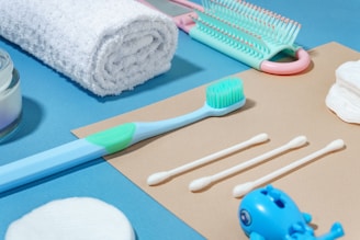 A neatly arranged collection of personal hygiene items on a two-toned background, including a rolled white towel, a toothbrush, a set of cotton swabs, a hairbrush, cotton rounds, and a small blue toy. The arrangement is aesthetically pleasing and conveys a sense of cleanliness and order.