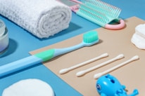 A neatly arranged collection of personal hygiene items on a two-toned background, including a rolled white towel, a toothbrush, a set of cotton swabs, a hairbrush, cotton rounds, and a small blue toy. The arrangement is aesthetically pleasing and conveys a sense of cleanliness and order.