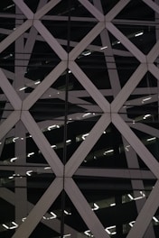 The image features an architectural facade with a geometric pattern created by intersecting diagonal lines forming diamond shapes. The structure appears modern, with a combination of glass and metal materials. The reflections and lighting within the structure add depth and complexity to the overall visual.
