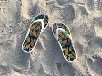 A pair of flip-flops with a pineapple pattern rests on sandy terrain, featuring footprints and textured sand around them.