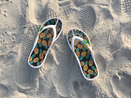 A pair of flip-flops with a pineapple pattern rests on sandy terrain, featuring footprints and textured sand around them.