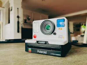 A vintage-style Polaroid OneStep 2 i-Type camera rests on a wooden surface. The camera features a large lens in the center, a red button, and a blue viewfinder on the right side. The background has a blurred interior of a room with soft lighting, including some out-of-focus wall decorations.