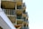 Multiple balconies with glass railings are stacked vertically on a modern apartment building. The structure's sharp lines and angles create an organized pattern. The facade consists of concrete and glass, with some brick elements visible. The sky is bright and clear, enhancing the architectural features.
