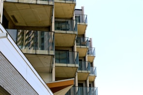 Multiple balconies with glass railings are stacked vertically on a modern apartment building. The structure's sharp lines and angles create an organized pattern. The facade consists of concrete and glass, with some brick elements visible. The sky is bright and clear, enhancing the architectural features.