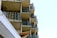 Multiple balconies with glass railings are stacked vertically on a modern apartment building. The structure's sharp lines and angles create an organized pattern. The facade consists of concrete and glass, with some brick elements visible. The sky is bright and clear, enhancing the architectural features.