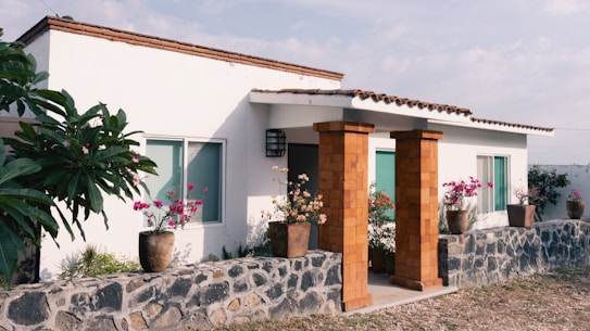 A modern white house with a flat roof and terracotta-colored columns. The front is adorned with potted plants featuring vibrant pink and red flowers. A stone wall borders the entrance path, enhancing the rustic charm.