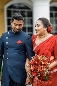 A couple dressed in elegant attire are standing closely together. The woman is wearing a red sari adorned with intricate patterns, holding a vibrant bouquet of red and orange flowers. The man is dressed in a formal dark blue outfit with metallic buttons and a red pocket square, matching the woman's attire. They appear to be in an outdoor setting, possibly outside a building with large windows and columns.