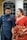 A couple dressed in elegant attire are standing closely together. The woman is wearing a red sari adorned with intricate patterns, holding a vibrant bouquet of red and orange flowers. The man is dressed in a formal dark blue outfit with metallic buttons and a red pocket square, matching the woman's attire. They appear to be in an outdoor setting, possibly outside a building with large windows and columns.