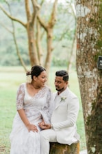 A couple dressed in wedding attire sits together on a tree stump in a lush, green outdoor setting. The woman wears a white lace gown with intricate details, and the man is in a white suit with a boutonniere. They are holding hands and looking fondly at each other, surrounded by trees and grass.