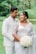 A couple is standing in a lush, green outdoor setting. They are wearing elegant attire; the woman is in a white saree with intricate lace details, holding a bouquet of white roses, while the man is dressed in a white suit. They appear to be in a joyful and intimate moment.