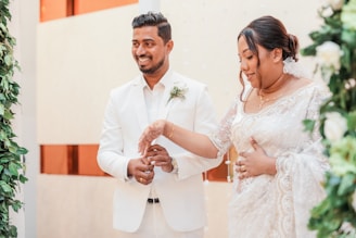 A couple dressed in elegant white attire stands together, surrounded by greenery and white floral decorations. The man is wearing a white suit with a boutonniere, while the woman is wearing a white lace dress. Both appear to be smiling and engaged in a ceremonial gesture.