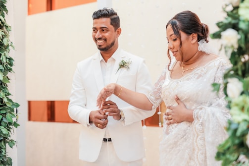 A couple dressed in elegant white attire stands together, surrounded by greenery and white floral decorations. The man is wearing a white suit with a boutonniere, while the woman is wearing a white lace dress. Both appear to be smiling and engaged in a ceremonial gesture.