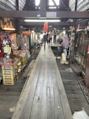 A wooden walkway leads through a rustic, indoor market area with various stalls selling souvenirs and local goods. The atmosphere is busy, with a few people visible in the distance. The market is housed in an older, wooden structure, adding a traditional charm to the setting.