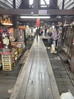 A wooden walkway leads through a rustic, indoor market area with various stalls selling souvenirs and local goods. The atmosphere is busy, with a few people visible in the distance. The market is housed in an older, wooden structure, adding a traditional charm to the setting.