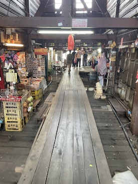 A wooden walkway leads through a rustic, indoor market area with various stalls selling souvenirs and local goods. The atmosphere is busy, with a few people visible in the distance. The market is housed in an older, wooden structure, adding a traditional charm to the setting.